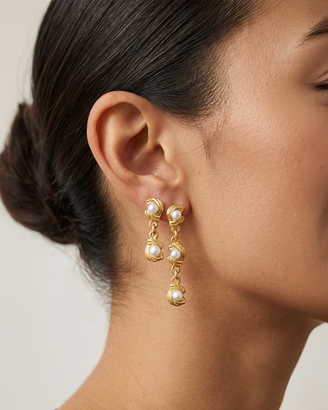Close-up of a person wearing gold earrings with pearl-like stones on a neutral background