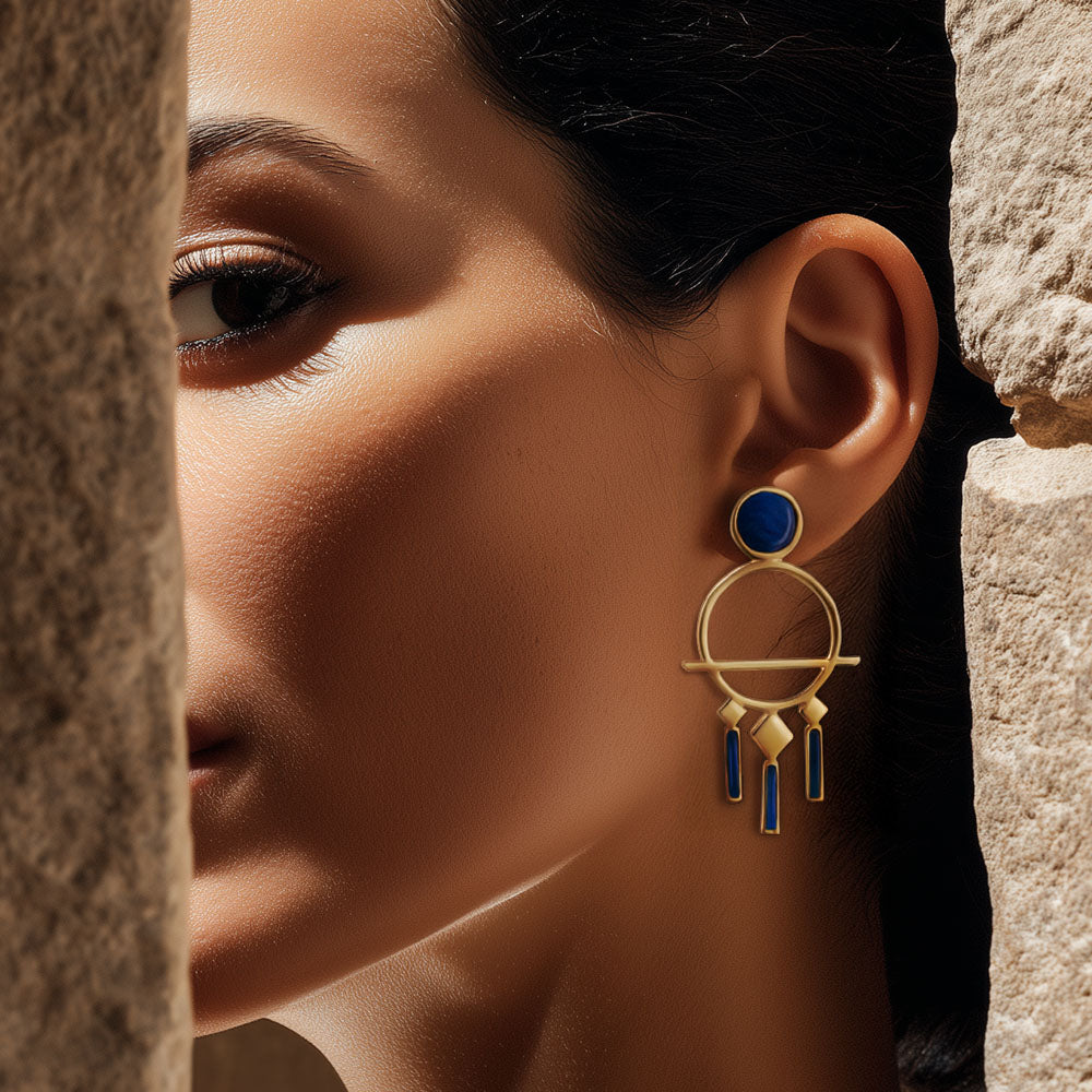 Close-up of a woman wearing a gold and blue earring against a stone wall, Gold Nefertiti profile earrings featuring a detailed textured headdress and a polished gold finish.